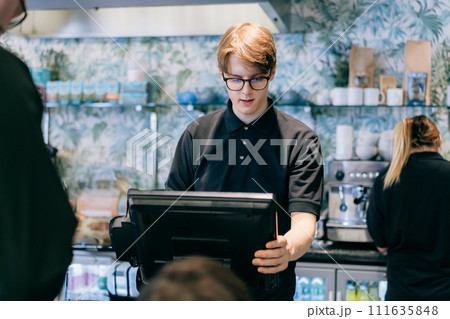 Young waiter serving customer at cash point in cafe. Man working with POS terminal. Cashier, barista checking for payment receipt. Hospitality, server and preparing a slip at the till in coffee shop. 111635848