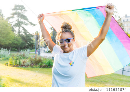 Mixed race woman raising a rainbow flag outdoor. Young lesbian activist smiling and holding flag symbol of social movement Lgbtq. Fight for equality, freedom and human rights. Celebrating Pride month. 111635874