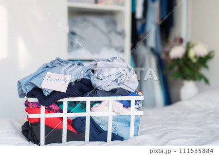A basket of selected clothes for donating to a Charity shop standing on the bed. Decluttering Clothes, Sorting, And Cleaning Up. Reuse, second-hand. Conscious consumer, sustainability. Selective focus 111635884