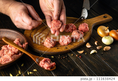 The chef hand holds a ground beef meatball ready for cooking. The concept of cooking cutlets on the kitchen table with aromatic spices The chef hand holds a ground beef meatball ready for cooking. The concept of cooking cutlets on the kitchen table with aromatic spices 111638424