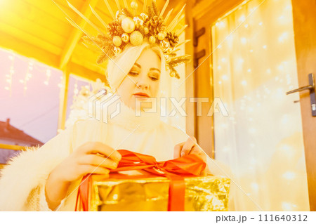 A woman in a white dress is holding a gold box with a red ribbon. She is wearing a crown on her head. The woman appears to be opening the gift box. 111640312