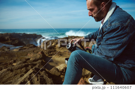 Caucasian handsome relaxed bearded man tourist pouring hot tea or coffee from thermos into a mug, resting by ocean 111640607