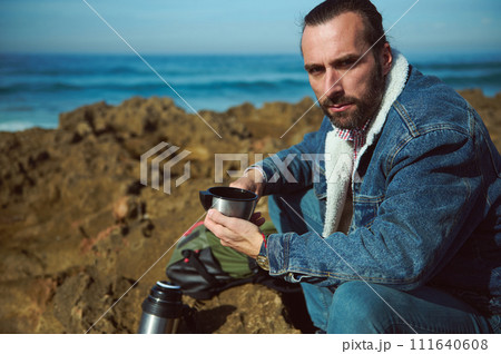 Lifestyle. Closeup view of a bearded man pouring hot water from a thermos, into a cup. The ocean in the background Lifestyle. Closeup view of a bearded man pouring hot water from a thermos, into a cup. The ocean in the background 111640608