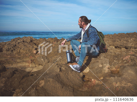 Relaxed young man traveler with backpack, sitting on the rocky cliff, enjoying the view of beautiful seascape 111640636