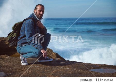 Bearded male traveler adventurer with backpack, sitting on the edge of a rocky cliff against breaking waves background Bearded male traveler adventurer with backpack, sitting on the edge of a rocky cliff against breaking waves background 111640638