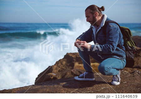 Handsome young male tourist checking his wristwatch, relaxing on the rocky cliff by sea with crushing waves 111640639