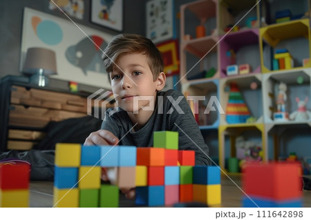 An eight-year-old boy assembles a house from multi-colored cubes while sitting on a wooden floor 111642898