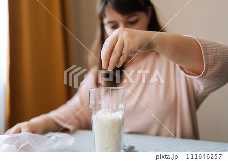 Close-up hands of focused female artisan pouring dry mixture or soy wax to glass for creating candle building mixture. Process of making handmade natural scented candle at workshop. Close-up hands of focused female artisan pouring dry mixture or soy wax to glass for creating candle building mixture. Process of making handmade natural scented candle at workshop. 111646257