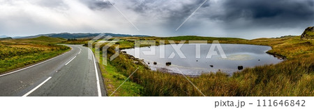 Narrow Road Beneath Small Pond In Wild Landscape Of Snowdonia National Park in Wales, United Kingdom Narrow Road Beneath Small Pond In Wild Landscape Of Snowdonia National Park in Wales, United Kingdom 111646842