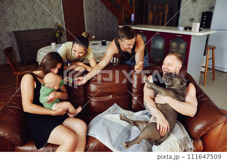 Young married couple with an American bully dog is sitting on sofa in living room while visiting young parents of baby girl. Young married couple with an American bully dog is sitting on sofa in living room while visiting young parents of baby girl. 111647509