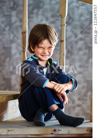 Dissatisfied grimace of little boy, 7 years old, sitting on step of wooden staircase leading to second floor of house in suburbs. Dissatisfied grimace of little boy, 7 years old, sitting on step of wooden staircase leading to second floor of house in suburbs. 111647557