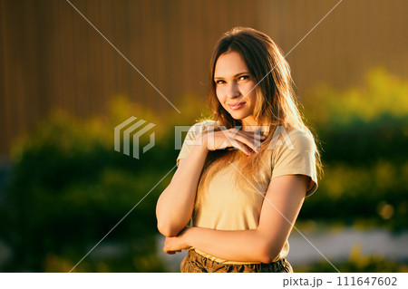 Portrait photography during golden hour, beautiful young Caucasian woman posing for photographer near rural house on summer evening, half length portrait of slender European female. 111647602