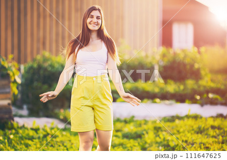 Portrait of woman in yellow Bermuda shorts, white female 26 years old posing for photographer near country house in summer before sunset. Portrait of woman in yellow Bermuda shorts, white female 26 years old posing for photographer near country house in summer before sunset. 111647625