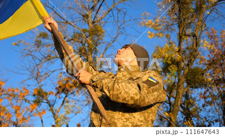 Male military in uniform waving flag of Ukraine at countryside. Young soldier of ukrainian army lifting blue-yellow banner as symbol of victory against russian aggression. Invasion resistance concept Male military in uniform waving flag of Ukraine at countryside. Young soldier of ukrainian army lifting blue-yellow banner as symbol of victory against russian aggression. Invasion resistance concept 111647643