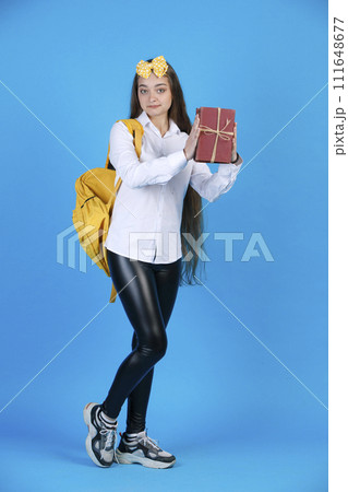 Pretty young woman in yellow bow raising bunch of books, while standing, shyly bending one leg in studio. Front view of cute female student demonstrating books, isolated on blue. Concept of posing. Pretty young woman in yellow bow raising bunch of books, while standing, shyly bending one leg in studio. Front view of cute female student demonstrating books, isolated on blue. Concept of posing. 111648677