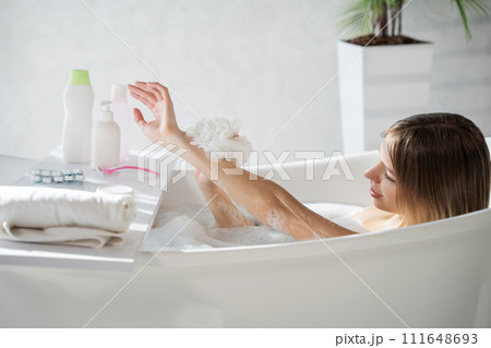 Happy caucasian lady washing arm with soapy sponge in bath. Side view of pretty young woman lather body with washcloth, while soaking in bathtub, with copy space. Concept of bathing, hygiene. 111648693