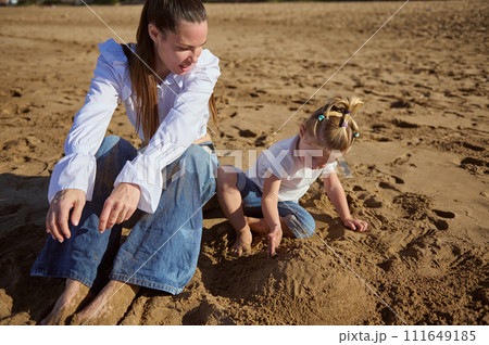 Happy family of a beautiful Caucasian single mother and daughter playing together on the beach, building sandy castle 111649185