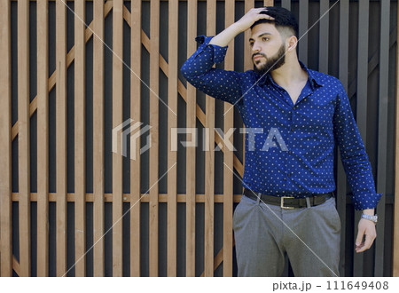 Front view of brunette asian gay standing, touching hair. Young businessman with beard wearing blue shirt and grey trousers, looking aside, thinking. Concept of urban life. Front view of brunette asian gay standing, touching hair. Young businessman with beard wearing blue shirt and grey trousers, looking aside, thinking. Concept of urban life. 111649408