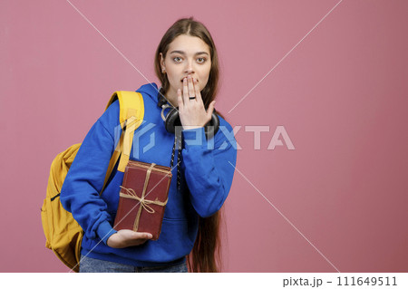 Front view of pretty schoolgirl standing, closing mouth by hand. Stylish female student holding bunch of old books, looking at camera. Isolated on pink studio background. Front view of pretty schoolgirl standing, closing mouth by hand. Stylish female student holding bunch of old books, looking at camera. Isolated on pink studio background. 111649511