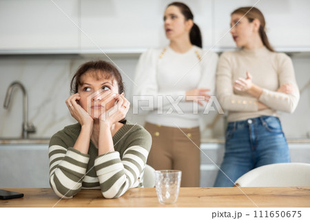 Upset middle-aged woman sitting at the kitchen table with her back to women quarreling to her 111650675
