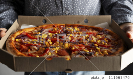 Caucasian male hands hold large open carton box with pepperoni pizza with sweetcorn front view Caucasian male hands hold large open carton box with pepperoni pizza with sweetcorn front view 111665476