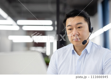 Serious Asian businessman engaging in customer service with headset in a contemporary workspace. 111666945