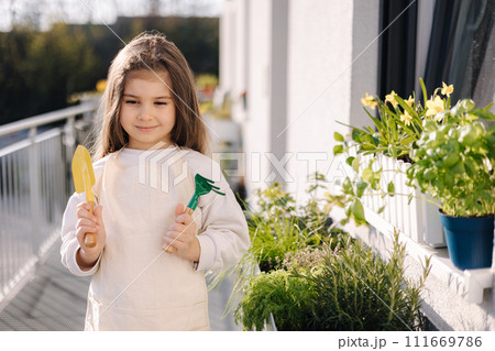Portrait of adorable little girl hold a small shovel and a rake and prepare for landing on balcony Portrait of adorable little girl hold a small shovel and a rake and prepare for landing on balcony 111669786