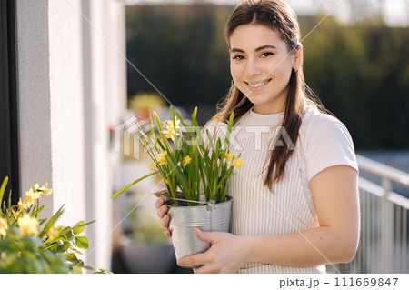 Beautiful female gardening outdoor. Happy woman hold metal bucket with daffodils. Flowerbed on balcony 111669847