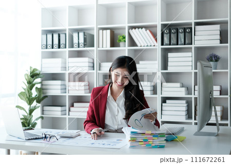 Beautiful asian businesswoman working on laptop and calculating with financial calculator from statistics data graphs, charts. Successful business results in modern office wear red shirt. 111672261