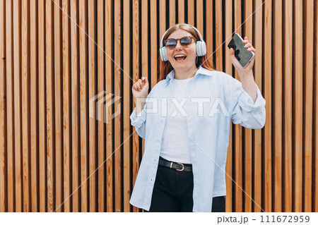 Closeup portrait of a dancing young woman listening to music through wireless earphones on wooden rustic background. Music lover enjoying music. Closeup portrait of a dancing young woman listening to music through wireless earphones on wooden rustic background. Music lover enjoying music. 111672959