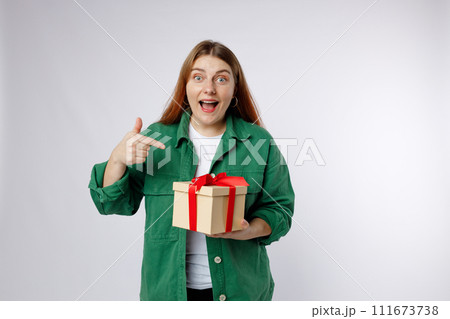 Portrait of a smiling casual woman holding present and pointing finger over white background. Happy woman receive gift, smiling excited. Valentine's Day Women's Day birthday holiday concept. Portrait of a smiling casual woman holding present and pointing finger over white background. Happy woman receive gift, smiling excited. Valentine's Day Women's Day birthday holiday concept. 111673738