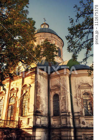 A weathered church with peeling paint stands amidst lush trees under a clear sky. The Church of St. John the Evangelist is an Orthodox church and an architectural monument in Nizhyn. A weathered church with peeling paint stands amidst lush trees under a clear sky. The Church of St. John the Evangelist is an Orthodox church and an architectural monument in Nizhyn. 111675378