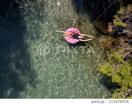 Aerial drone view of young woman with open arms in swimsuit lying and relaxing on big pink donut float swims on water surface with transparent bottom. Summer holiday vacation at exotic resort Aerial drone view of young woman with open arms in swimsuit lying and relaxing on big pink donut float swims on water surface with transparent bottom. Summer holiday vacation at exotic resort 111676018