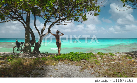 Woman enjoy beach bicycle stop, tropical island landscape. Slim girl tourist in bikini stand under tree on sea coast, relax after ride. Outdoor lifestyle travel on summer holiday vacation. Back view 111678162