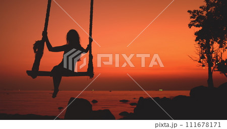 Happy woman silhouette swinging in tropical sunset sitting on swing with scenic view on orange sky, sea waves. Outdoor lifestyle travel on summer holiday vacation, enjoy freedom, have fun. Happy woman silhouette swinging in tropical sunset sitting on swing with scenic view on orange sky, sea waves. Outdoor lifestyle travel on summer holiday vacation, enjoy freedom, have fun. 111678171