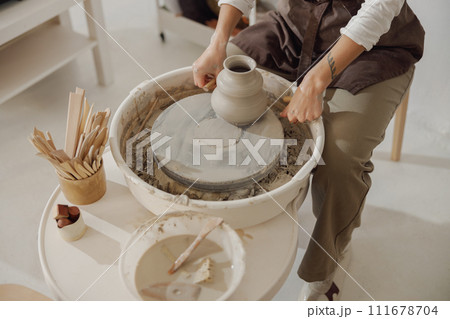 Female potter in apron making shape of clay vase on spinning pottery tool in ceramic workshop Female potter in apron making shape of clay vase on spinning pottery tool in ceramic workshop 111678704