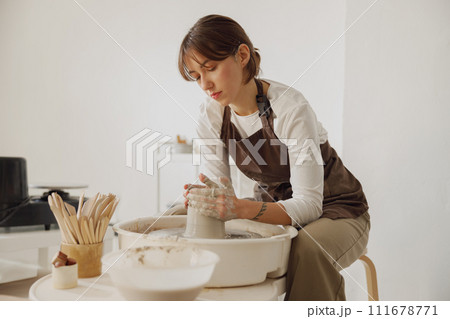 Concentrated female artisan in apron sitting on bench with pottery wheel and making clay pot 111678771