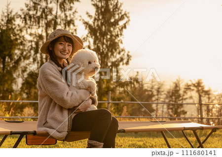 秋冬のキャンプ場で愛犬を抱くキャンパーのアジア人女性(ビジョンフリーゼ) 秋冬のキャンプ場で愛犬を抱くキャンパーのアジア人女性(ビジョンフリーゼ) 111680141