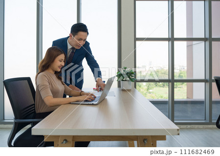 Young asian woman in casual wear typing on laptop computer keyboard. Her boss in suit and tie, stand pointing at the screen, discussing work or collaborating on a project. 111682469