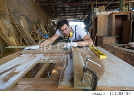 A man is focused on carving wood, using tools to shape it. Wood shavings are scattered around his workbench. 111682480