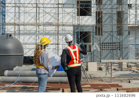 Two construction workers are standing together in front of a large building site with helmet and safety gear. Day time work safety checks. Work environment at the site of housing projects. Two construction workers are standing together in front of a large building site with helmet and safety gear. Day time work safety checks. Work environment at the site of housing projects. 111682696