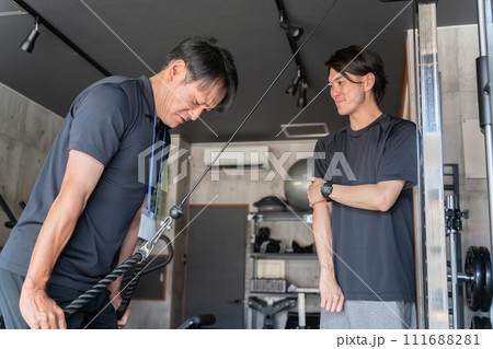 パーソナルジムで会員にトレーニングの見本を見せるトレーナーの男性(腕トレ) パーソナルジムで会員にトレーニングの見本を見せるトレーナーの男性(腕トレ) 111688281
