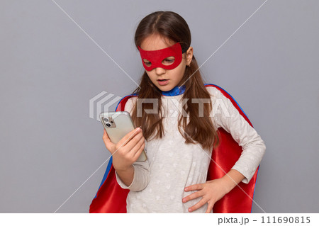 Concentrated little girl wearing superhero costume and mask isolated over gray background reading text message on smartphone holding hand on hip having displeased facial expression Concentrated little girl wearing superhero costume and mask isolated over gray background reading text message on smartphone holding hand on hip having displeased facial expression 111690815