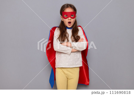 Shocked little girl wearing superhero costume and mask isolated over gray background standing with crossed arms saying omg keeping mouth widely opened 111690818