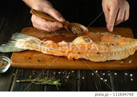 The chef adds coriander with a spoon to the raw fish fillet on the kitchen table. Concept of preparing fish herring in restaurant or public house kitchen 111694798