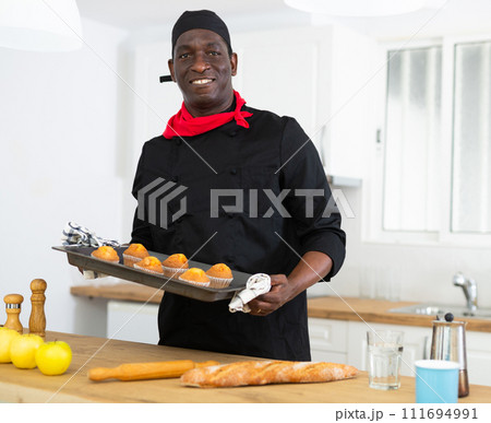 Proffesional man in black chef uniform holding sheet pan with just baked cupcakes Proffesional man in black chef uniform holding sheet pan with just baked cupcakes 111694991