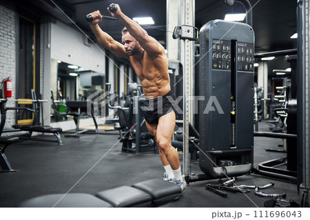 Middle-aged Caucasian man training in crossover machine indoors. Side view of bearded crossfit male athlete doing pull-downs, lifting metal plates of cable crossover machine in gym. Crossfit concept. Middle-aged Caucasian man training in crossover machine indoors. Side view of bearded crossfit male athlete doing pull-downs, lifting metal plates of cable crossover machine in gym. Crossfit concept. 111696043
