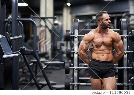 Pensive shirtless crossfit athlete leaning on palf rack barbell platform, having rest after training. Front view of strong, muscular guy looking away, while exercising in gym. Concept of sport. 111696055
