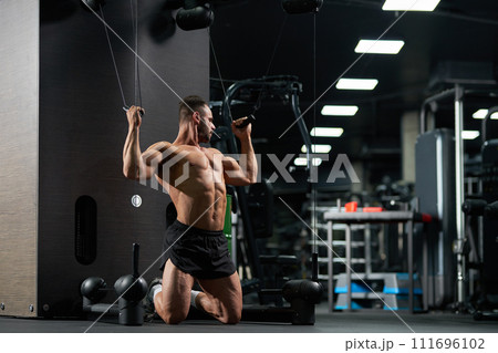 Strong sportsman kneeling, while doing exercise for biceps in gym. Full body of muscular male athlete wearing black shorts, pulling cables of training apparatus indoors. Concept of sport, workout. Strong sportsman kneeling, while doing exercise for biceps in gym. Full body of muscular male athlete wearing black shorts, pulling cables of training apparatus indoors. Concept of sport, workout. 111696102