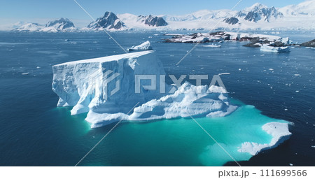 Melting iceberg in Antarctica in sunny day. Huge glacier in turquoise sea water, snow covered mountains in background. Aerial drone flight panorama. Global warming, climate change. Explore South Pole 111699566